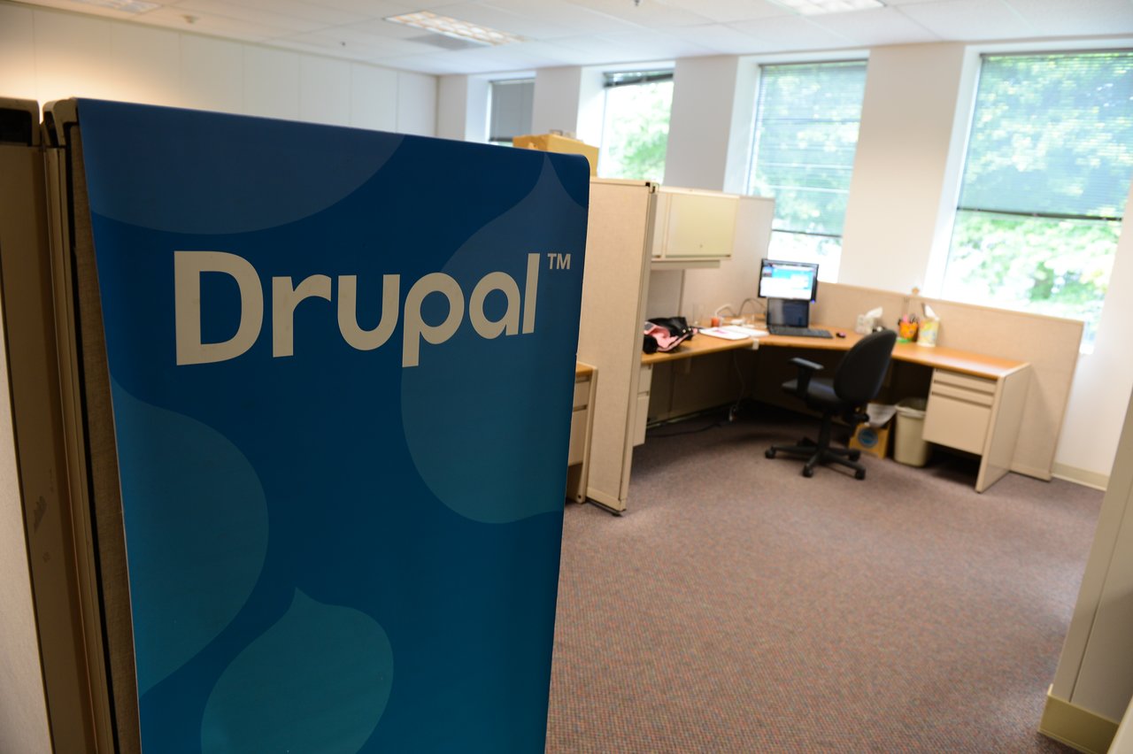 A Drupal-branded sign in the foreground with an office workspace, desk, and computer in the background.