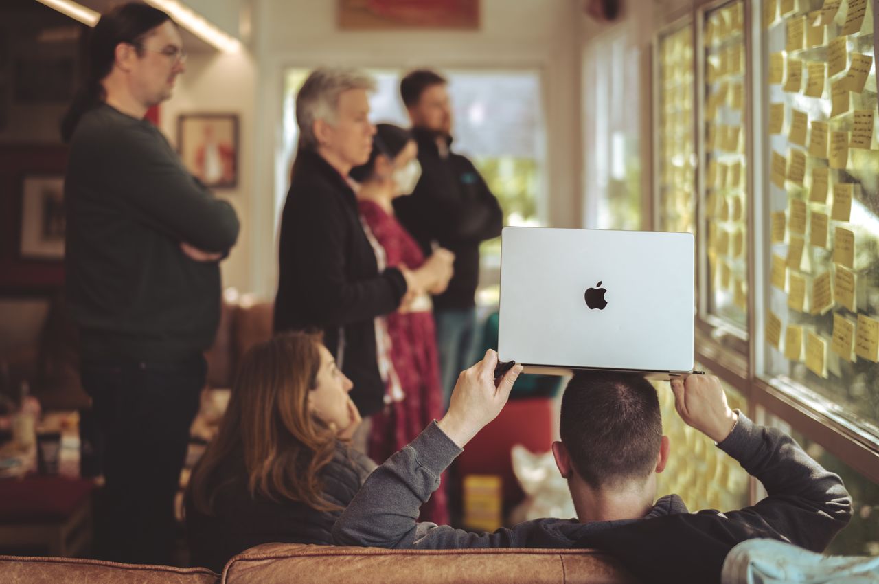 A man sits on a sofa with a laptop on his head, while a group of people gather around sticky notes in the background, working together.