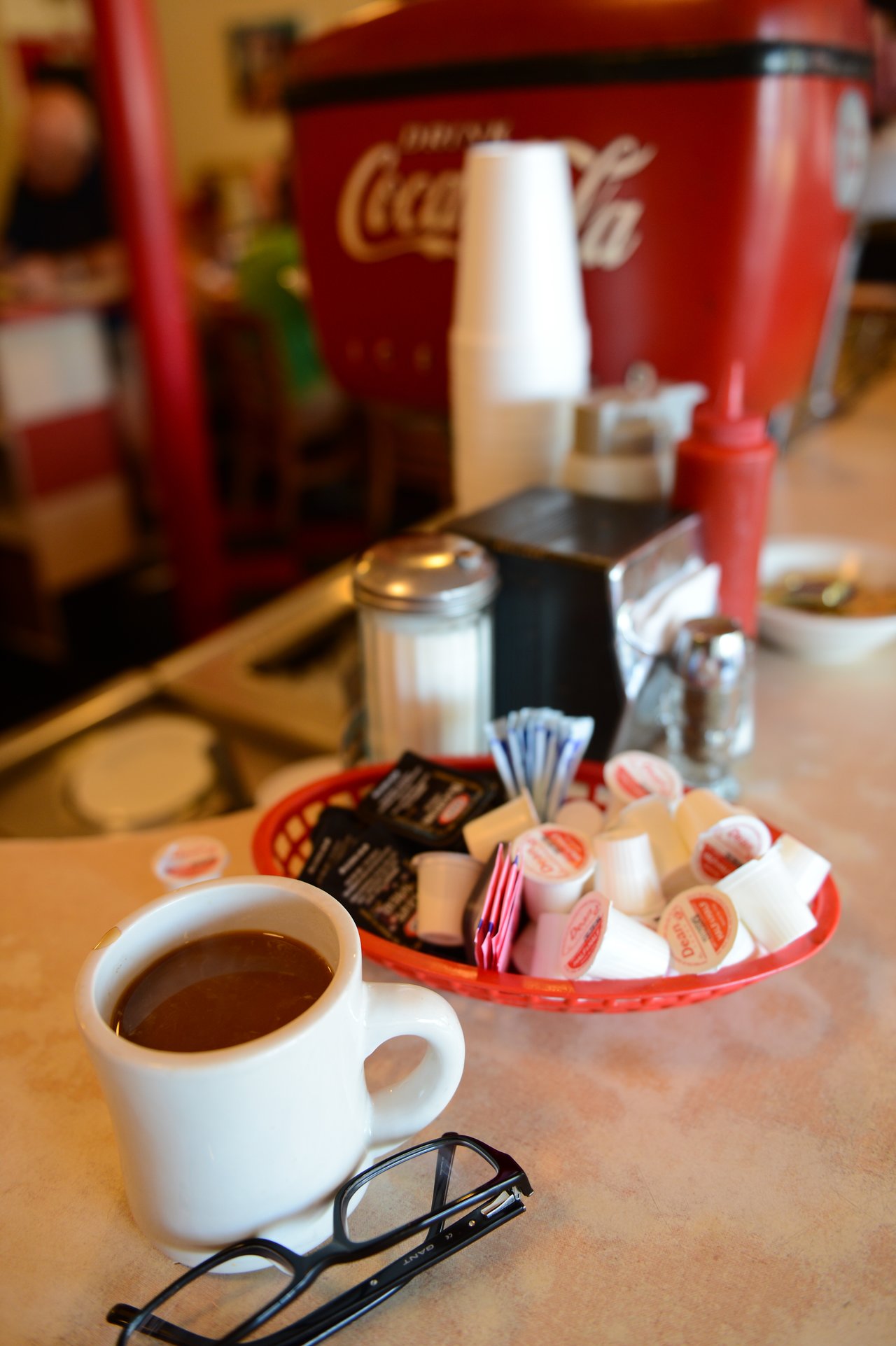 A cup of coffee sits on a diner counter beside a pair of glasses and a basket of creamers.