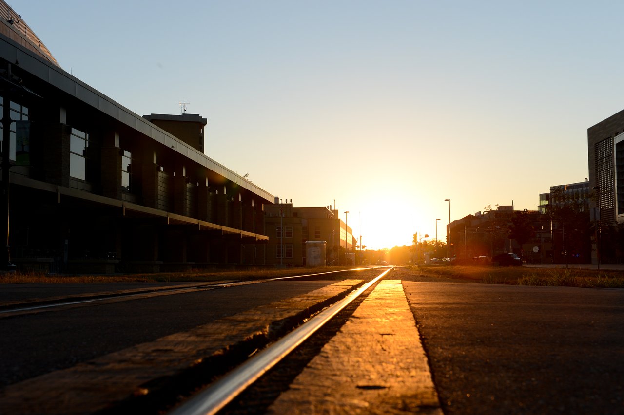 Low-angle view of railroad tracks leading into the sunset, surrounded by modern buildings and streetlights.