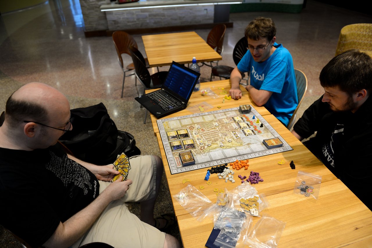 Three people play a strategy board game at a table, with a laptop open nearby.