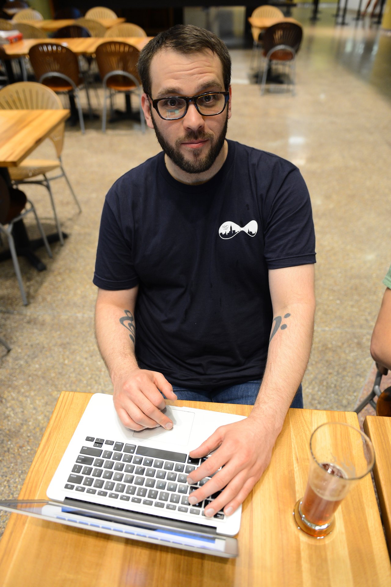 A person in a black Drupal t-shirt works on a laptop at a table with a drink nearby.