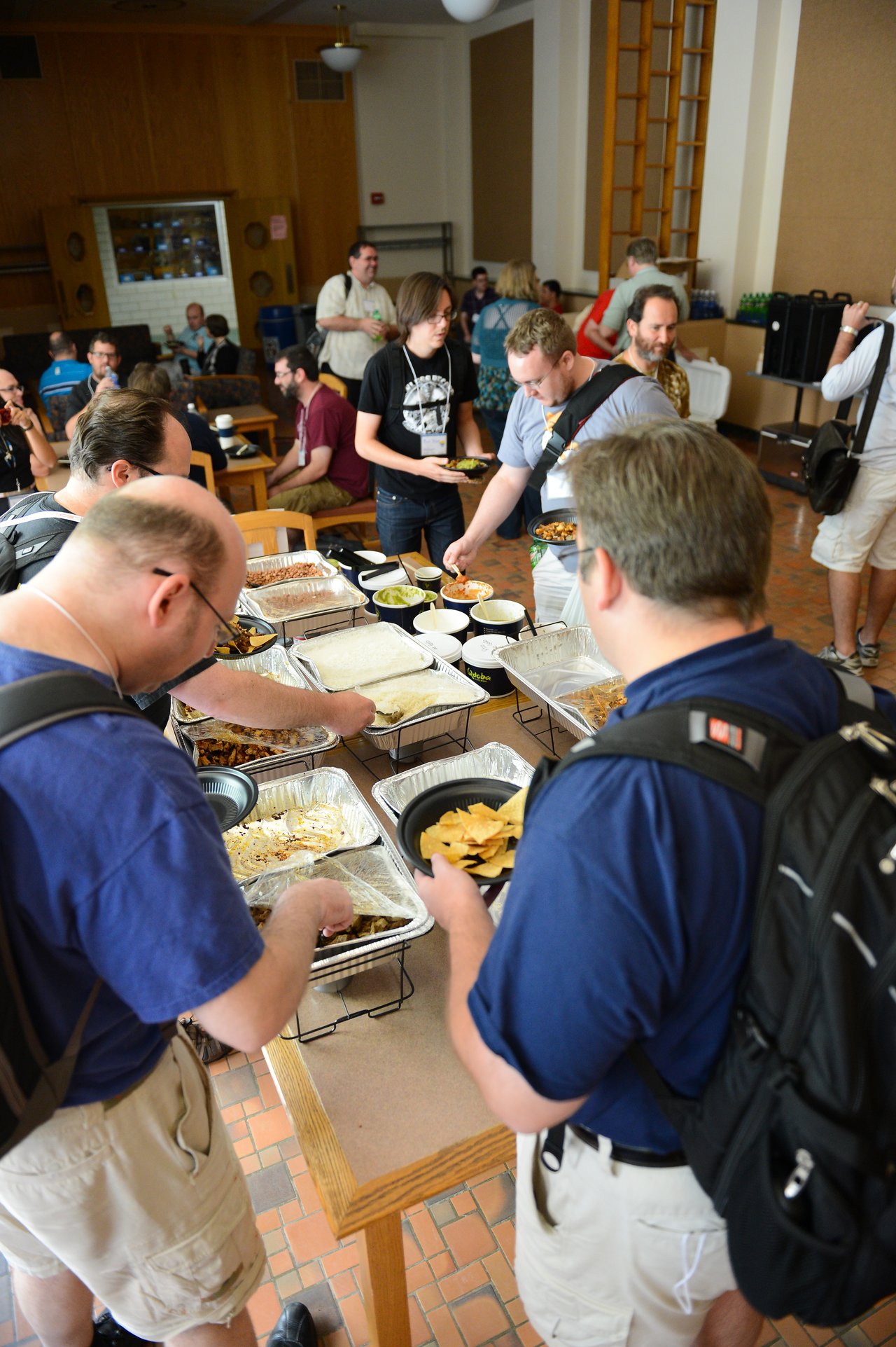 People serving themselves food from a buffet table at a gathering, with others socializing in the background.