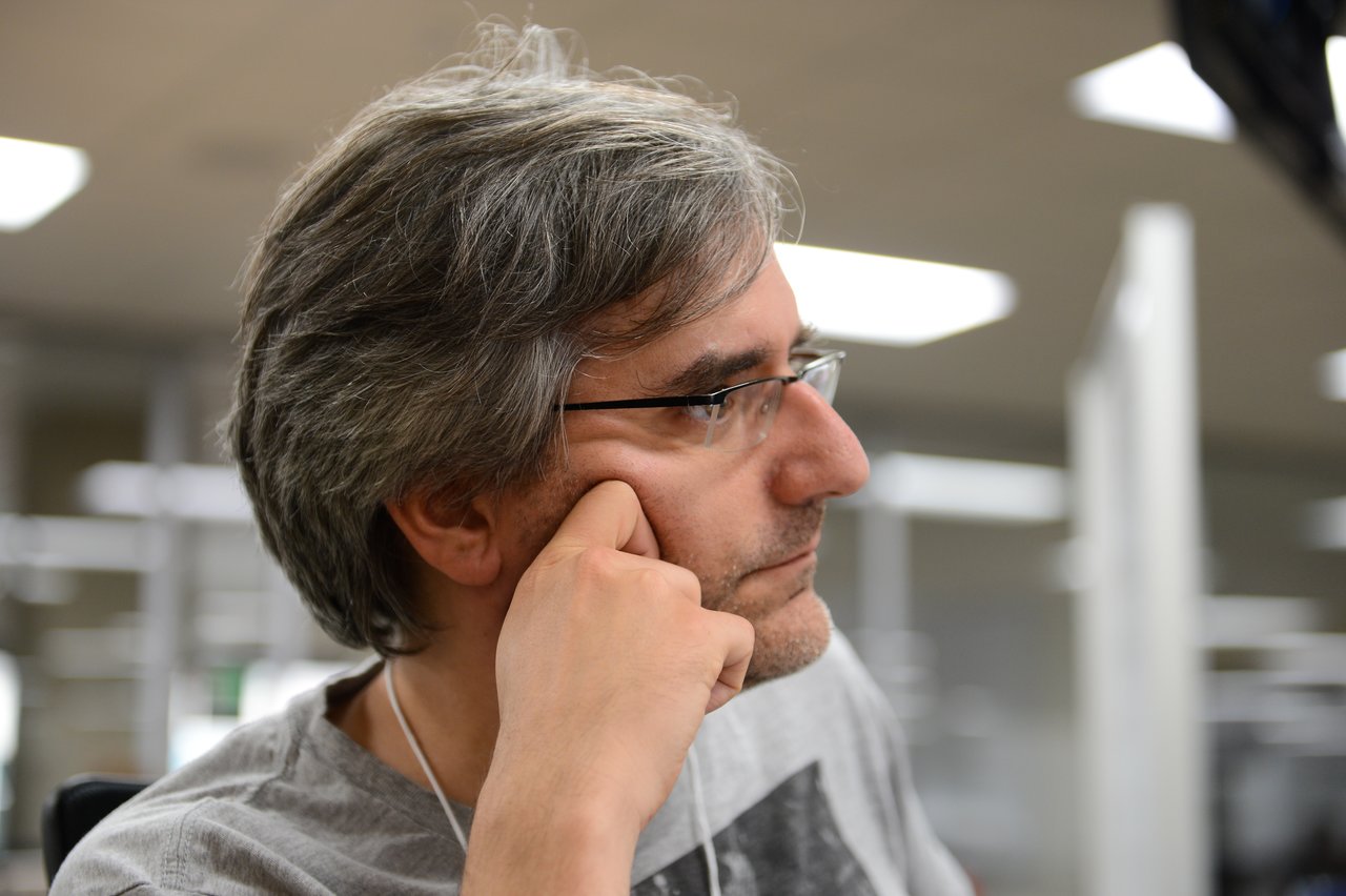 A person with glasses and gray hair sits, resting their face on their hand, appearing focused.
