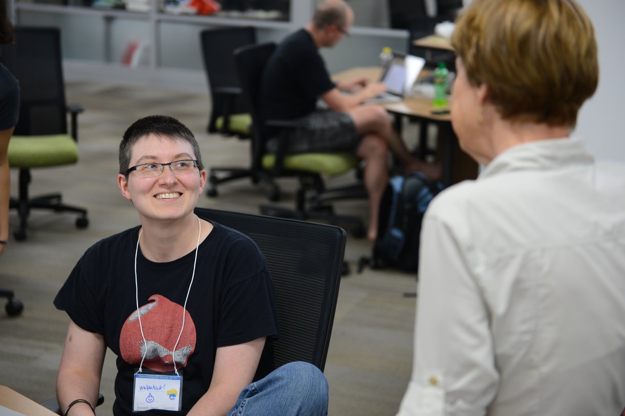 Two people engaged in conversation at a Drupal developer sprint, with others working on laptops in the background.