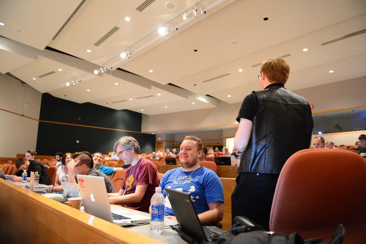 A group of people in a conference room work on laptops while someone stands and speaks to them.