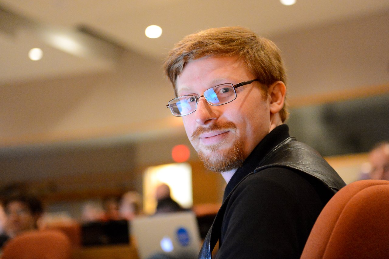 A person with glasses and a beard looks toward the camera while seated in a conference room.