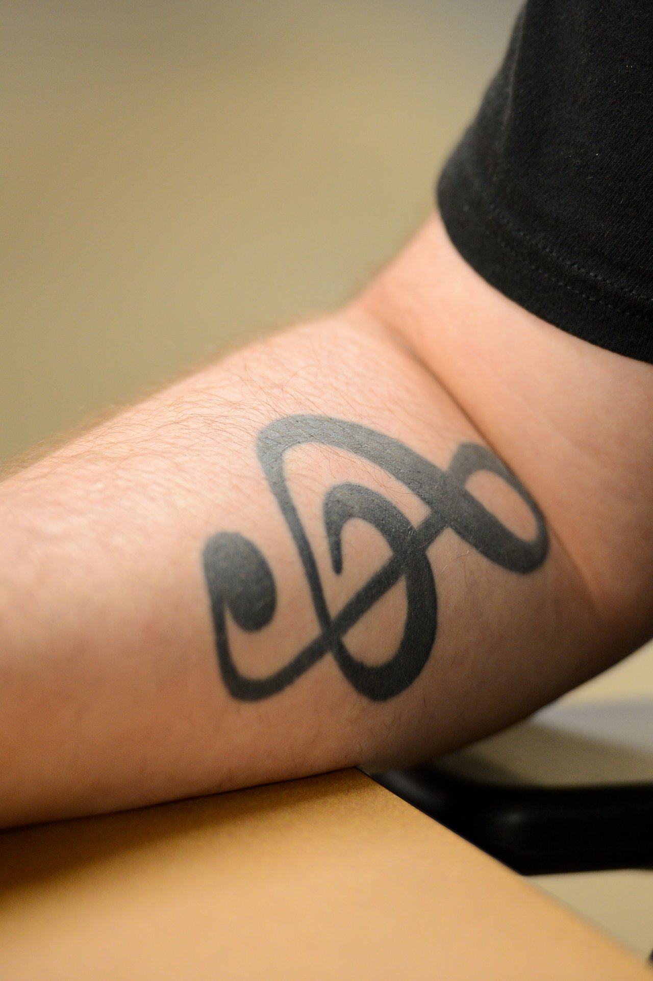 A close-up of a person's forearm resting on a table, showing a black tattoo of a treble clef.