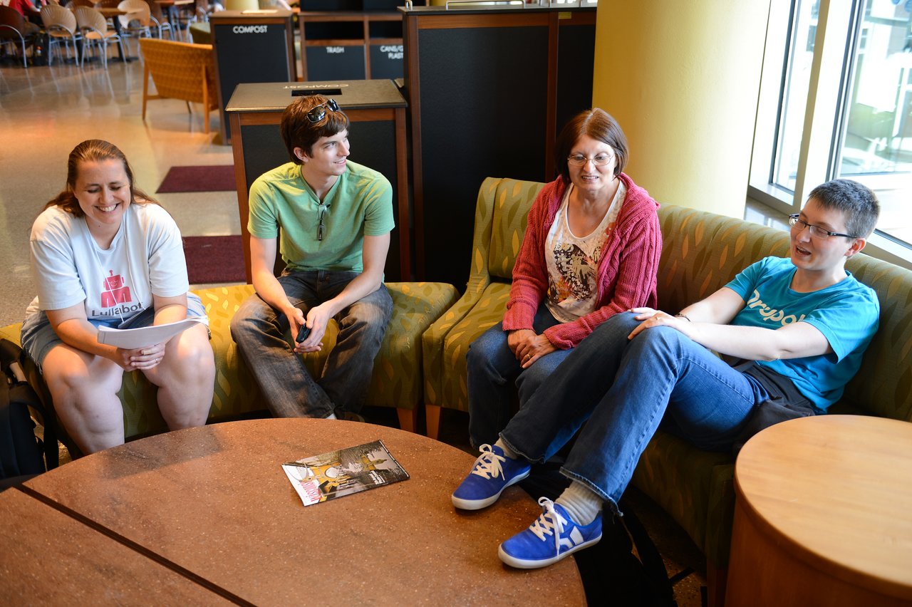 Four people sit on couches in a casual discussion, with one holding papers and another gesturing while speaking.