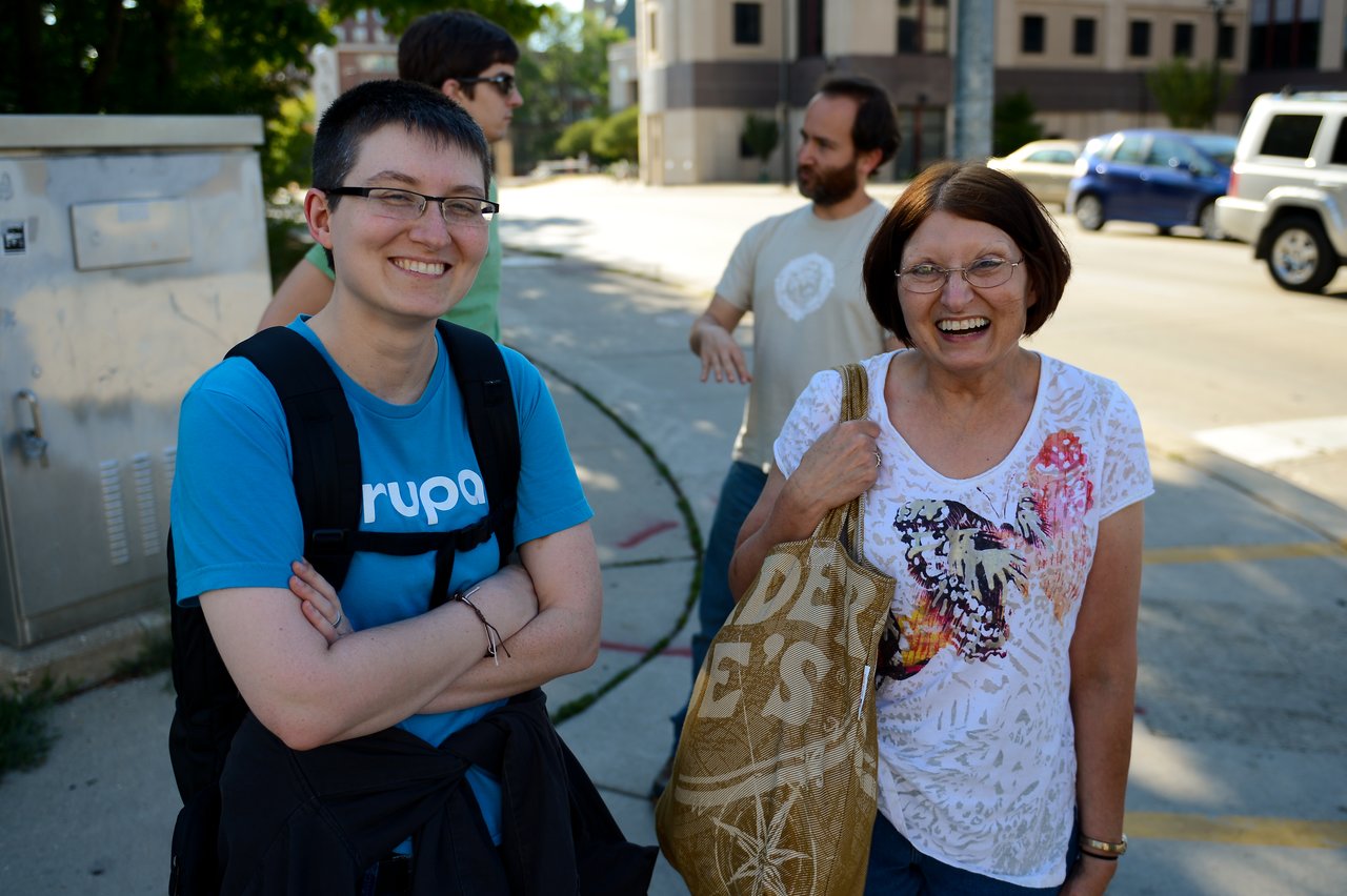 Two people smiling outdoors, one wearing a Drupal shirt and backpack, the other holding a reusable shopping bag.