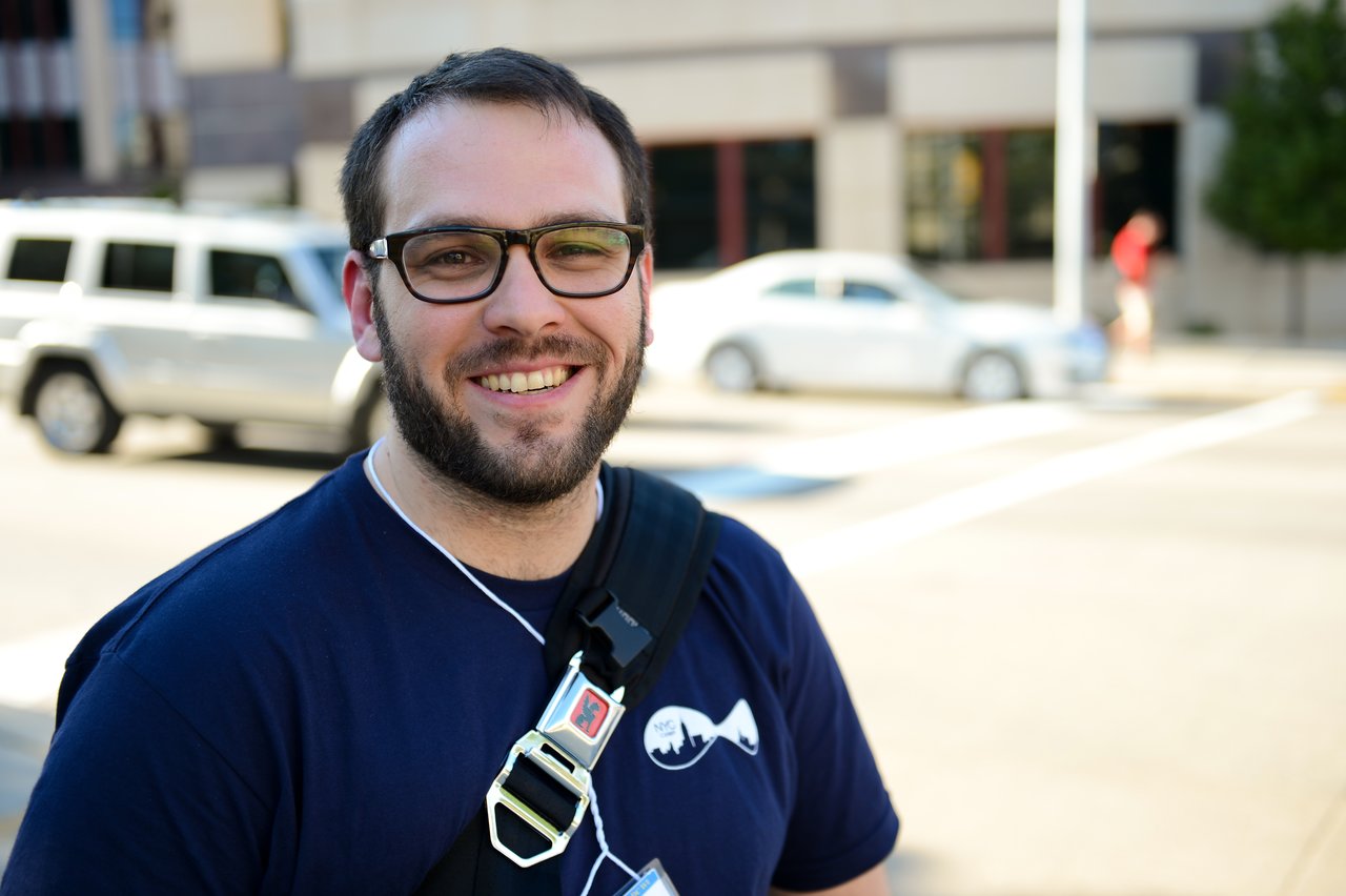 A smiling person wearing glasses and a conference badge stands outdoors with cars and buildings in the background.