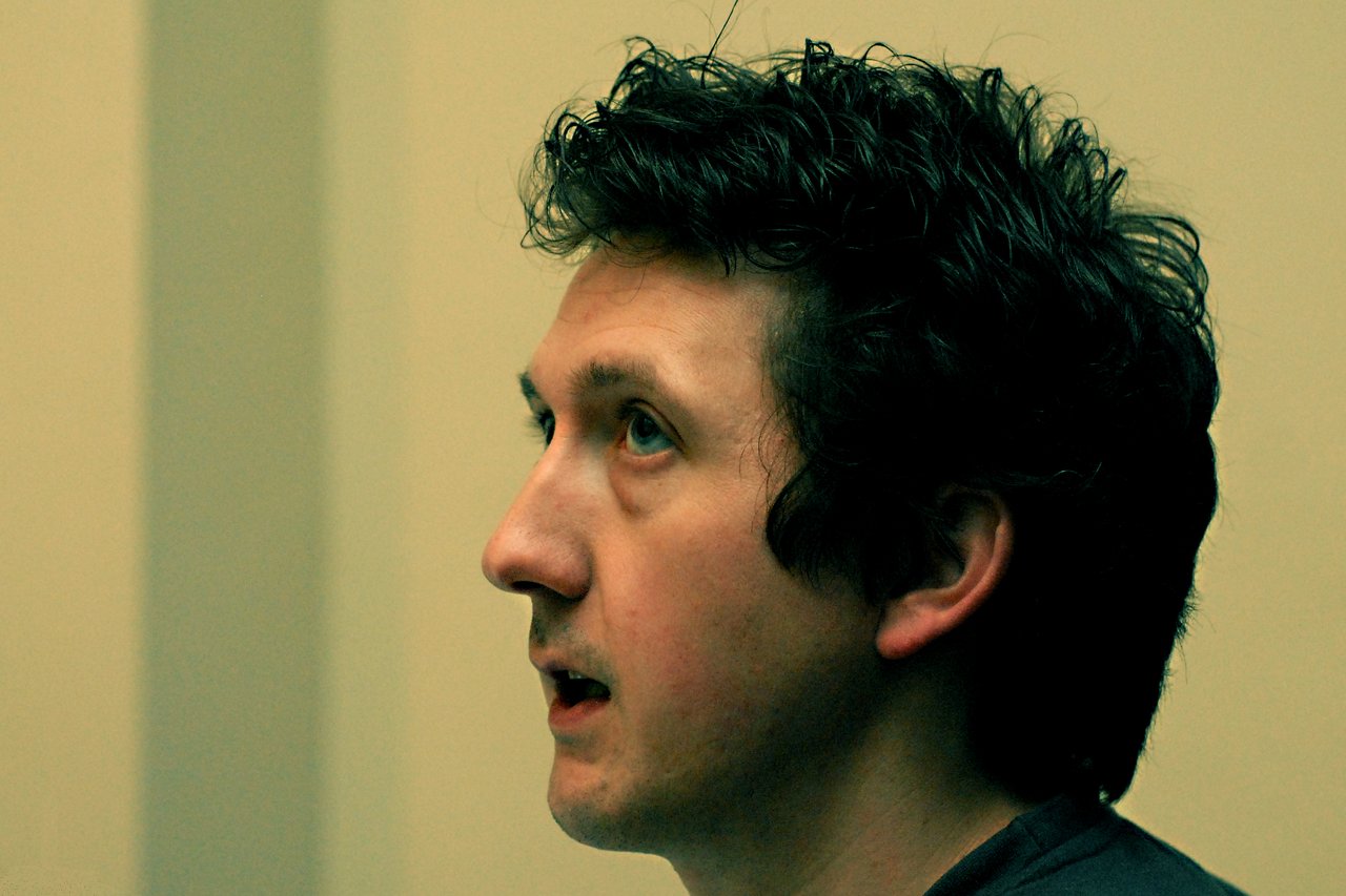 A man with dark curly hair speaks while looking ahead in an indoor setting.