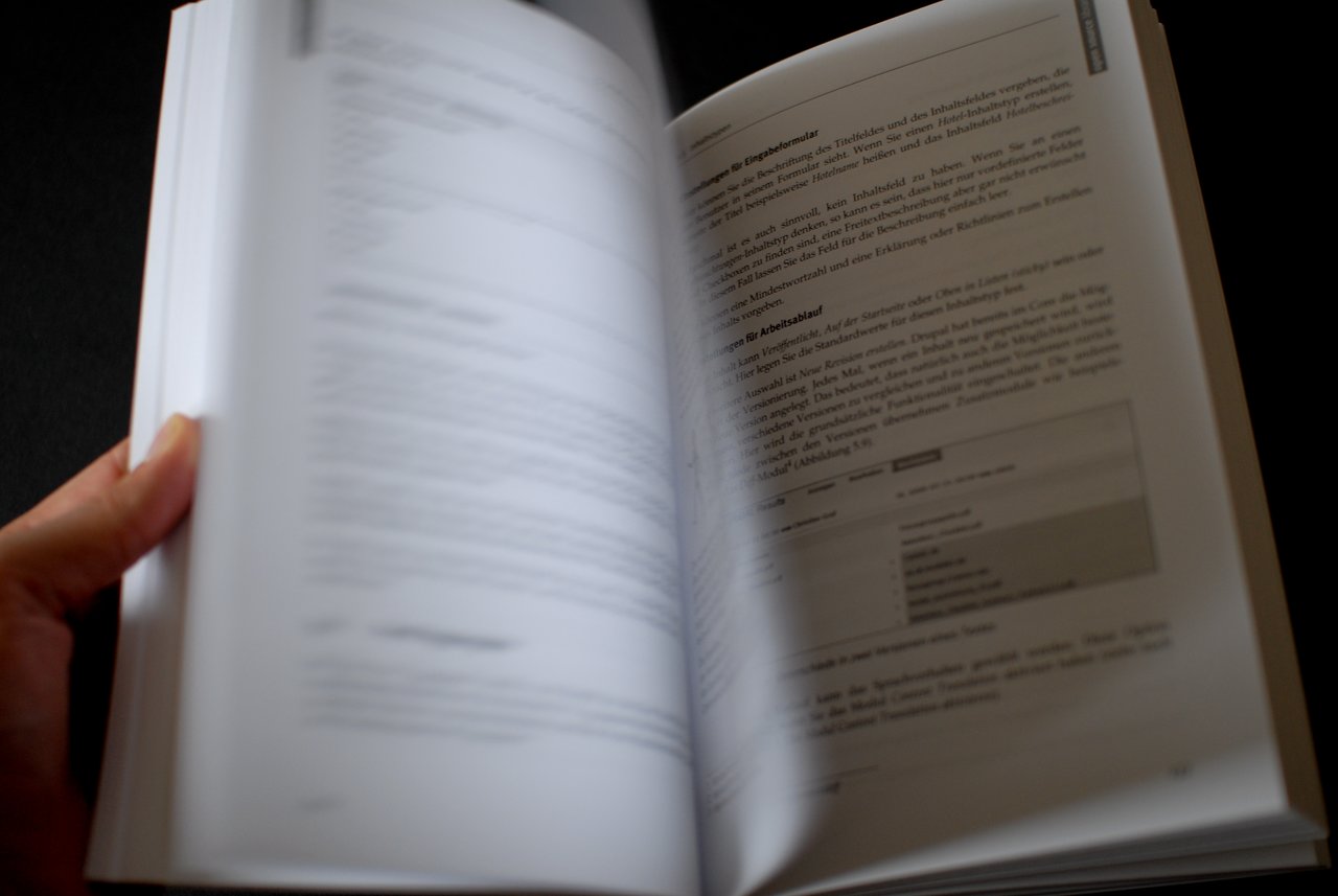A person flips through a book about Drupal 6, with blurred pages showing text and a screenshot.