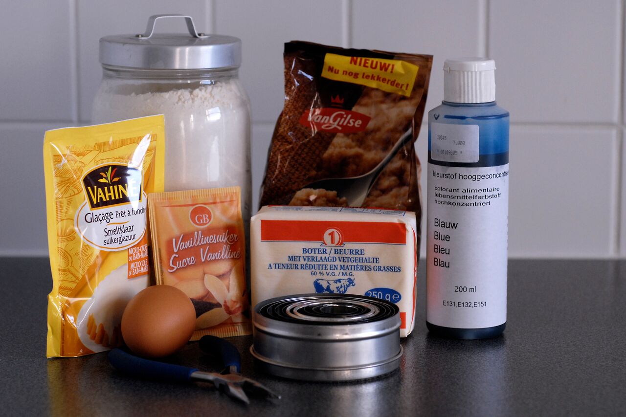 Baking ingredients and tools, including flour, sugar, butter, an egg, and food coloring, arranged on a countertop.