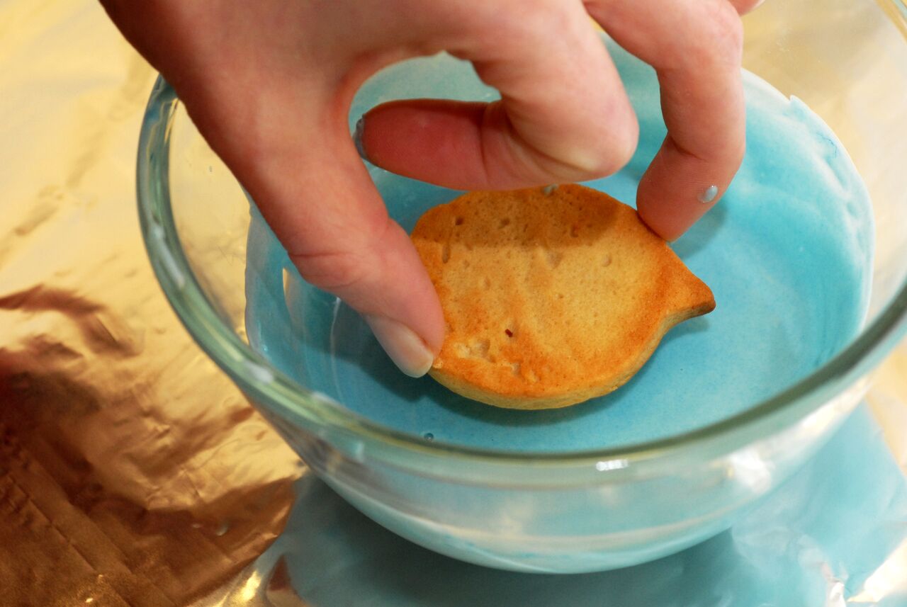 A hand dips a fish-shaped cookie into a bowl of blue frosting.