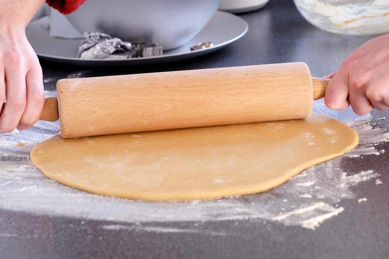 Hands rolling out cookie dough with a wooden rolling pin on a floured countertop.