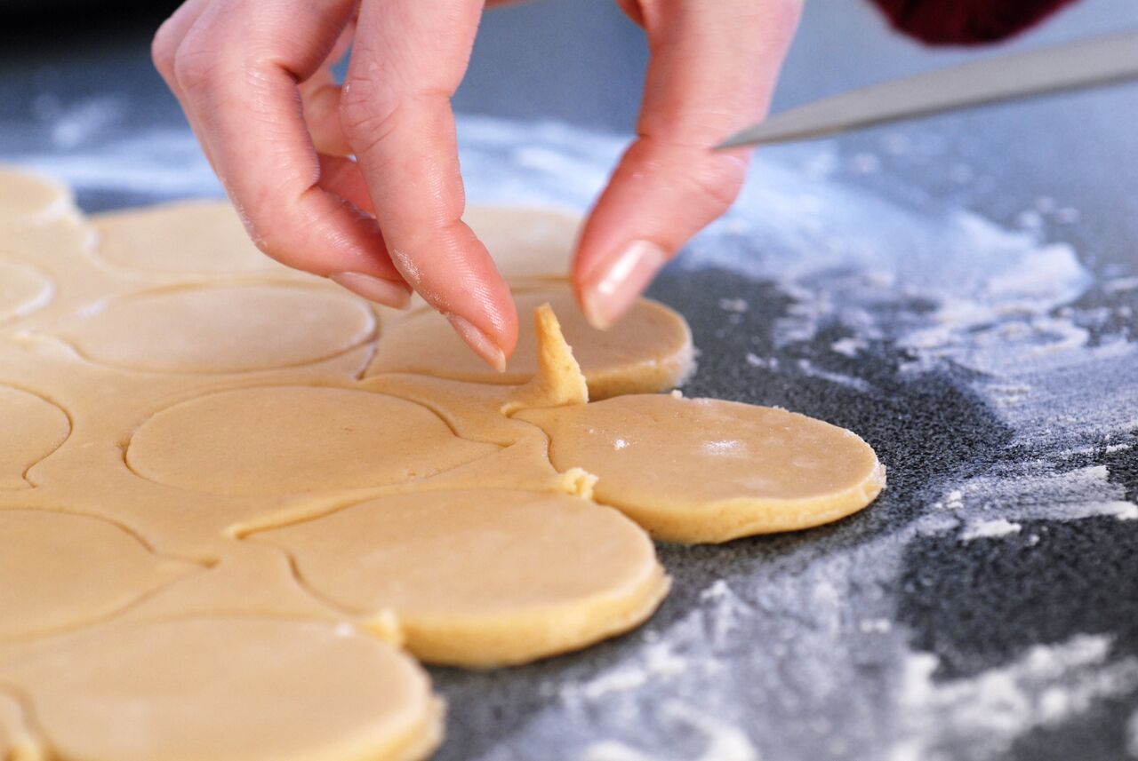 A person carefully lifts a small piece of cookie dough from a larger sheet on a floured surface.