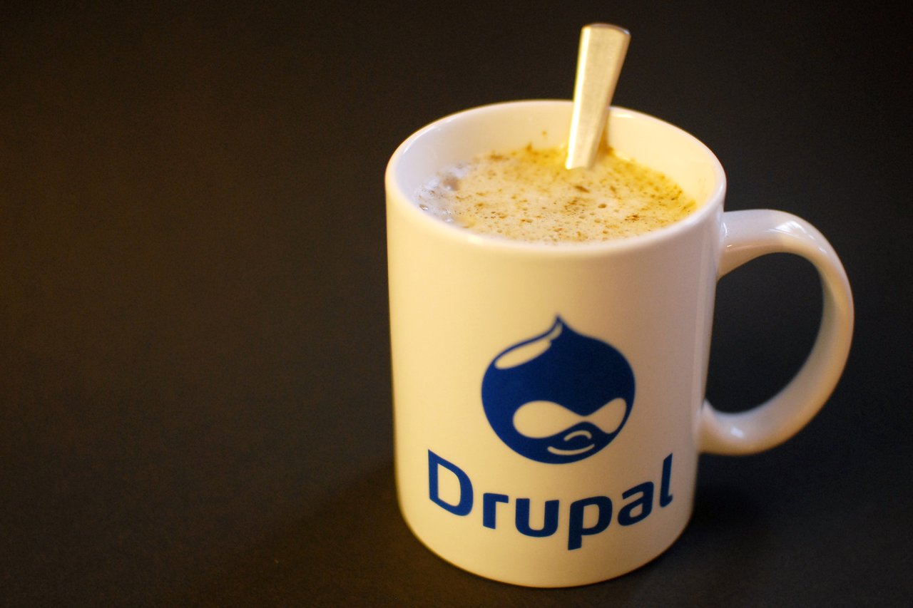 A white mug with the Drupal logo, filled with a frothy drink, with a spoon inside.