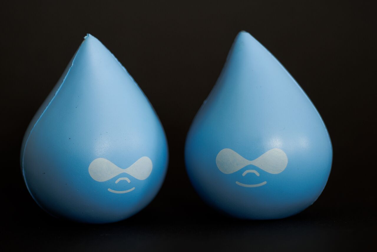 Two blue Druplicon stress balls with smiling faces, placed side by side against a dark background.
