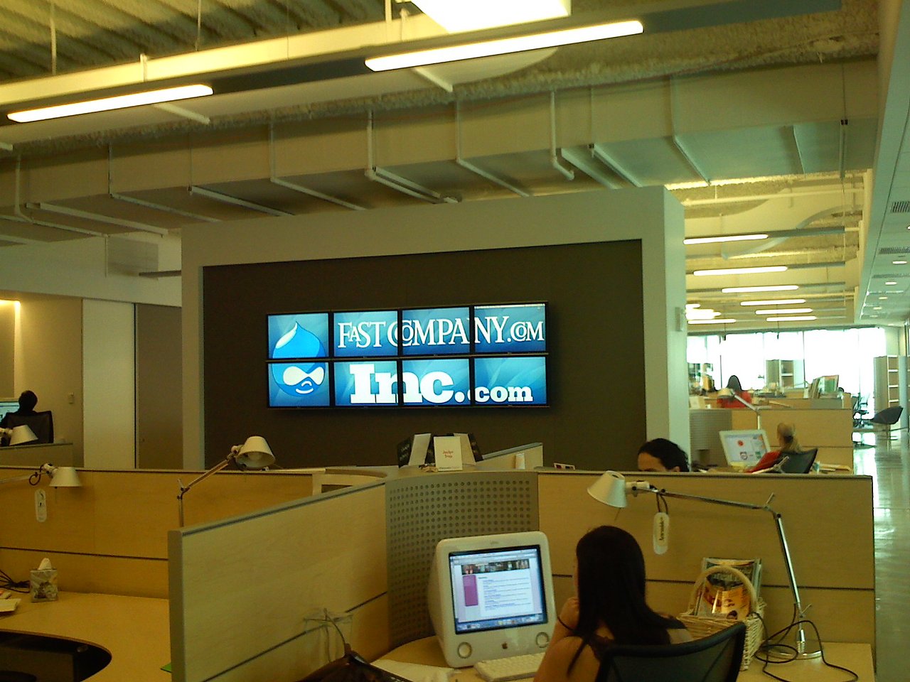An open office with employees working at desks, and a large screen displaying "FastCompany.