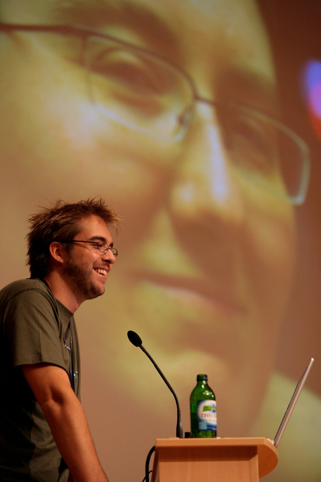 A speaker at a podium smiles while presenting, with a large projected face behind them.