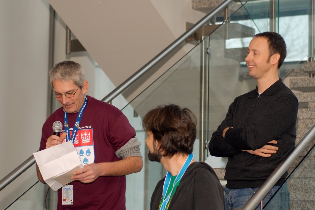 A man in a maroon shirt reads from a paper while speaking into a microphone, with two others listening.