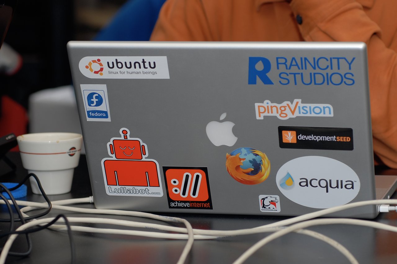 A laptop covered in various tech and Drupal-related stickers, including Ubuntu, Acquia, Lullabot, and Firefox.
