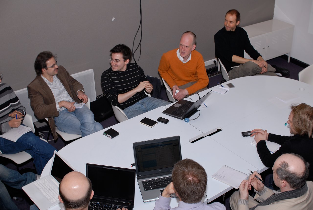 A group of people sitting around a table, discussing and using laptops during a Drupalcamp event.