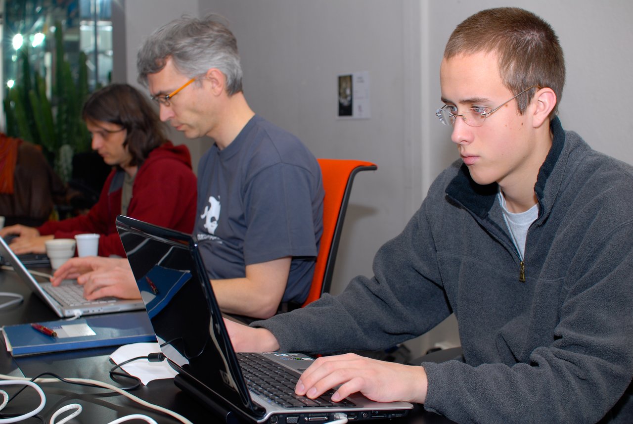 A young man in glasses and a gray fleece types on a laptop at a DrupalCamp Paris 2008 event.
