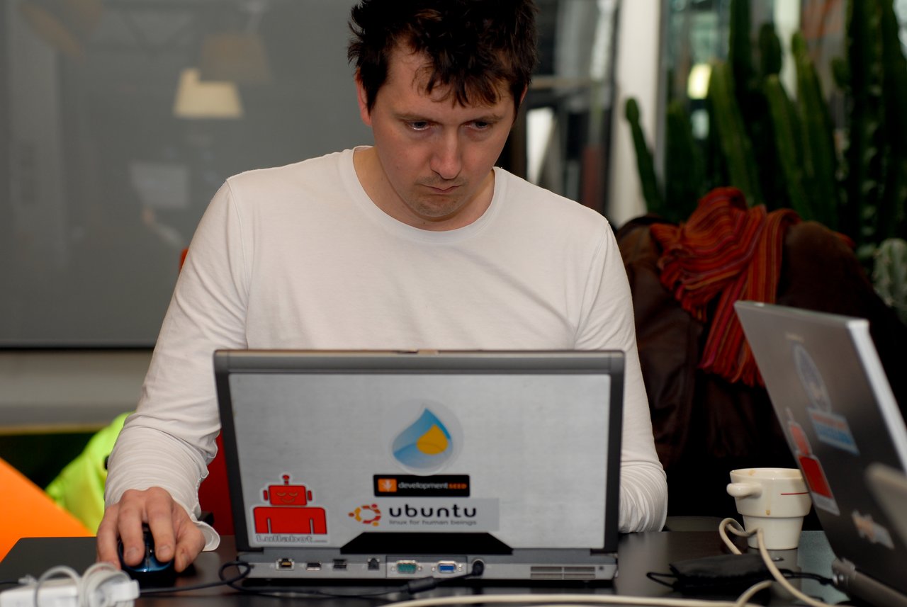 A man in a white shirt is focused on his laptop, using a mouse at a workspace.