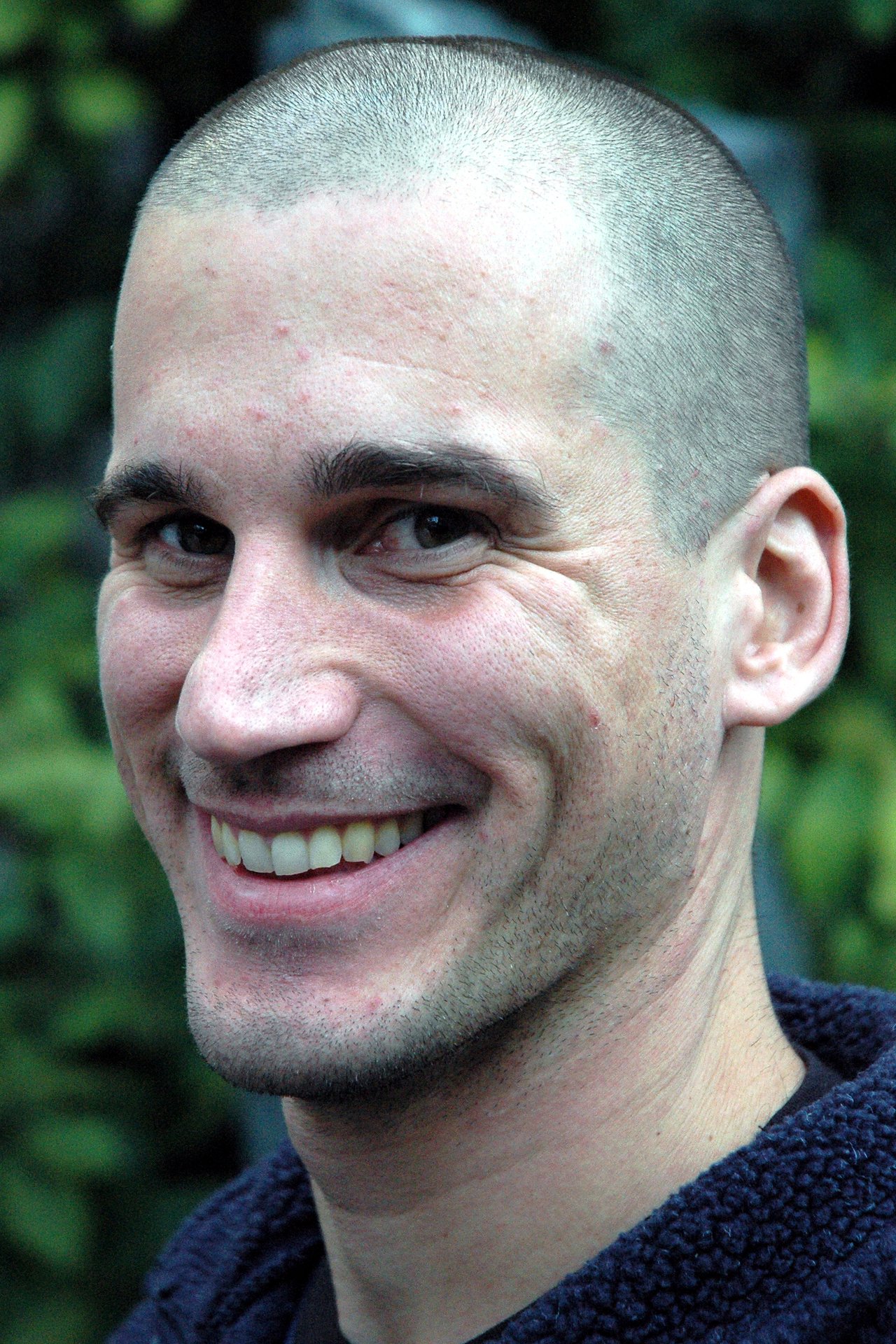 A person with a shaved head and a dark jacket smiles at the camera with a friendly expression.