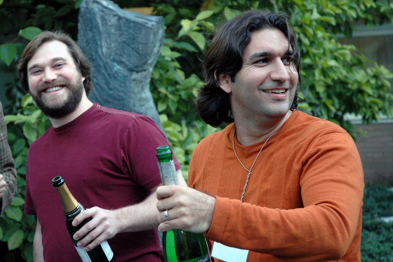 Two smiling people holding champagne bottles outdoors, appearing to celebrate or enjoy a gathering.