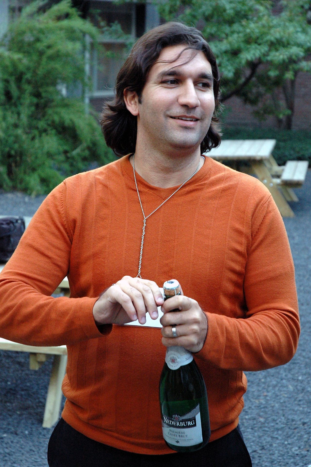 A person in an orange sweater is opening a bottle of sparkling wine while smiling and looking to the side.