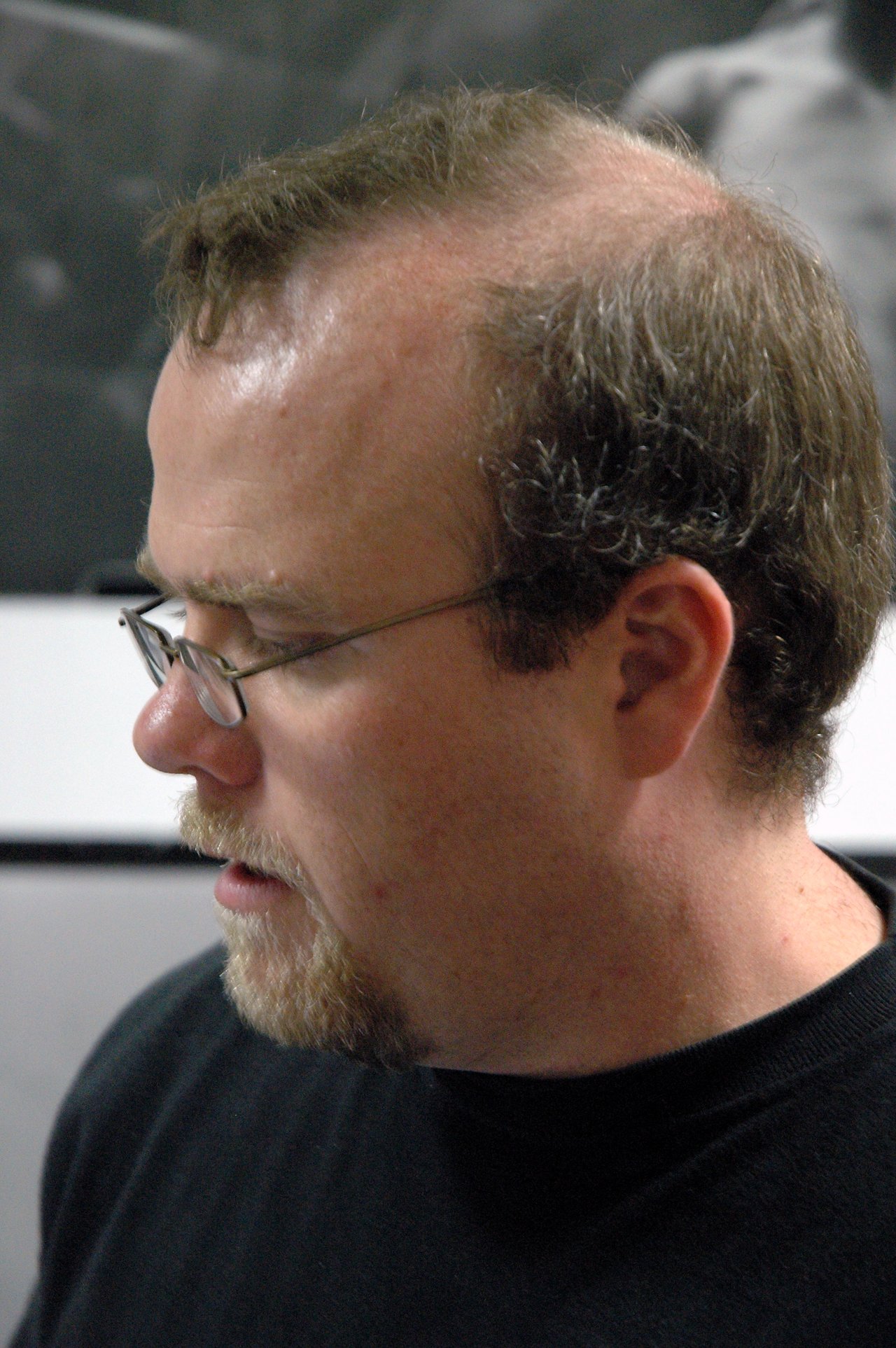 A man with glasses and a beard looks downward, appearing focused.