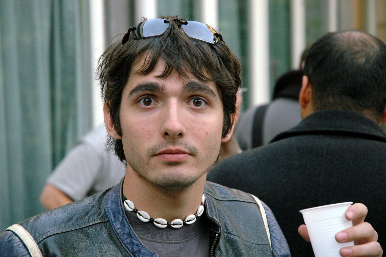 A person with sunglasses on their head and a shell necklace looks directly at the camera, holding a cup.