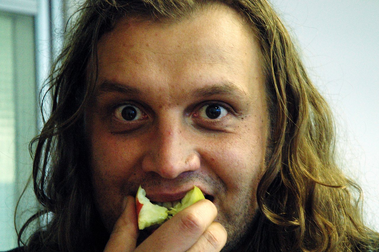 A person with long hair takes a bite of an apple while looking directly at the camera.