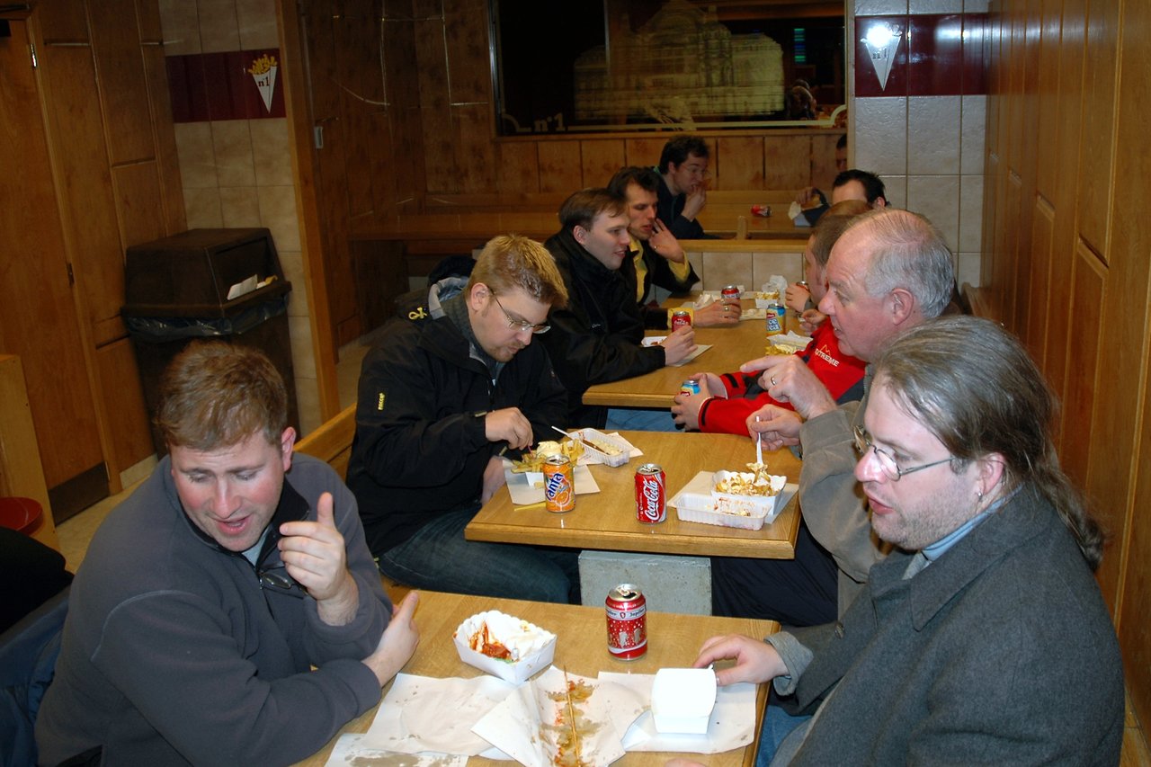 A group of people sitting at tables in a casual restaurant, eating Belgian fries and drinking canned soda.