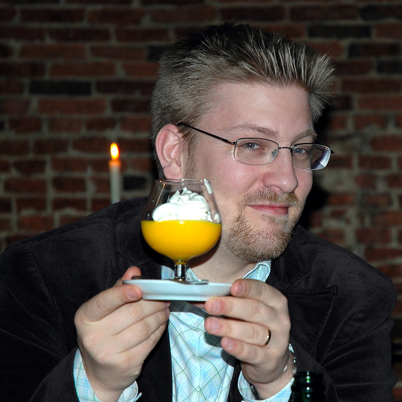 A man with glasses holds a glass of yellow dessert with whipped cream, smiling at the camera.
