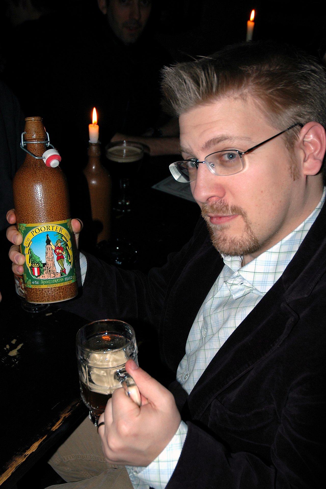 A man in glasses holds a Belgian beer bottle and a glass of beer in a dimly lit setting.