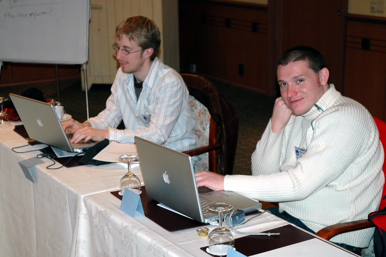 Two people sitting at a table with laptops, one typing and the other smiling at the camera.
