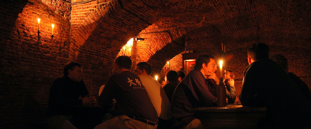 People sit at wooden tables in a dimly lit brick-walled room, talking by candlelight.