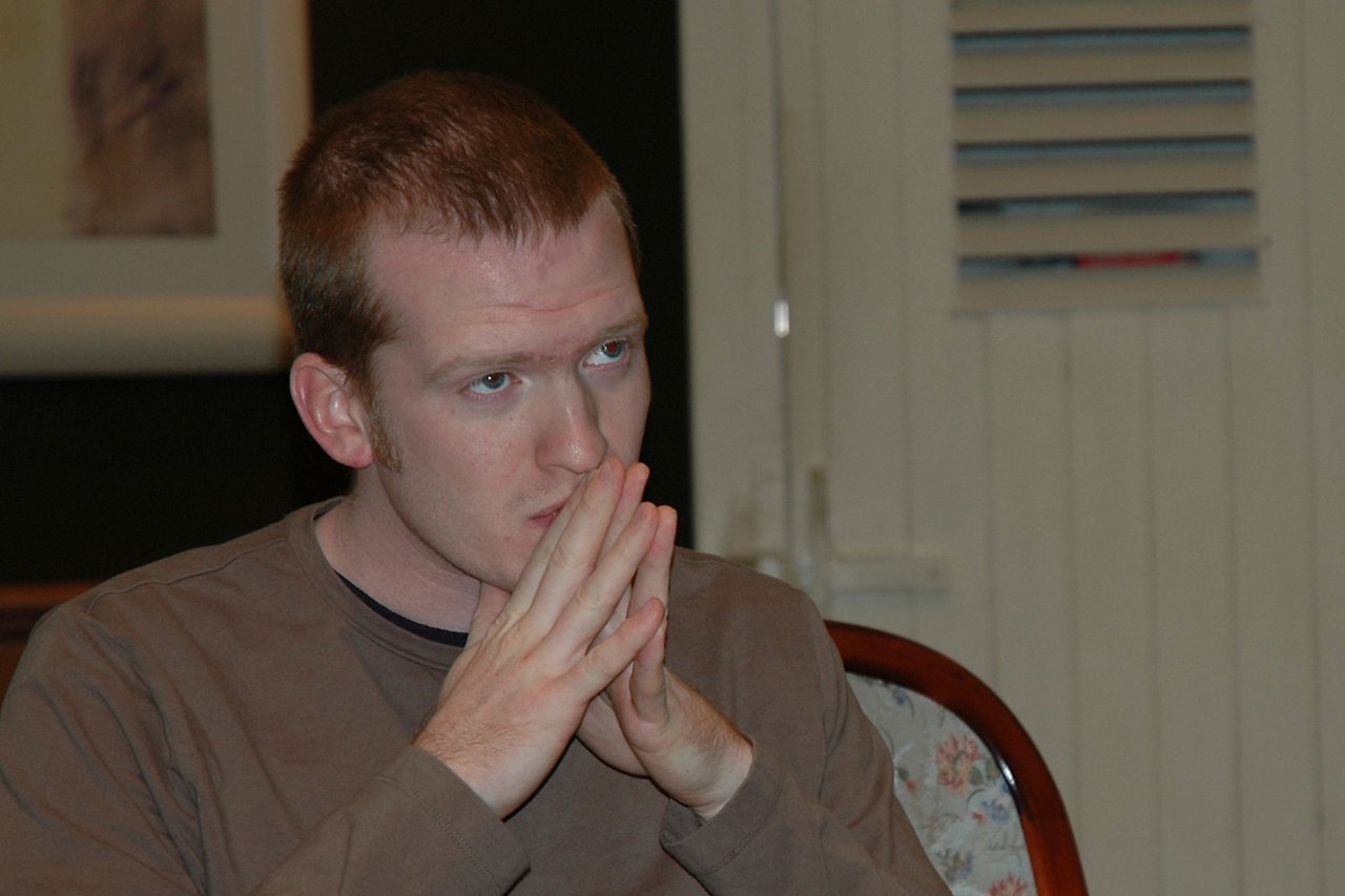 A person sits with hands pressed together, looking focused or deep in thought.