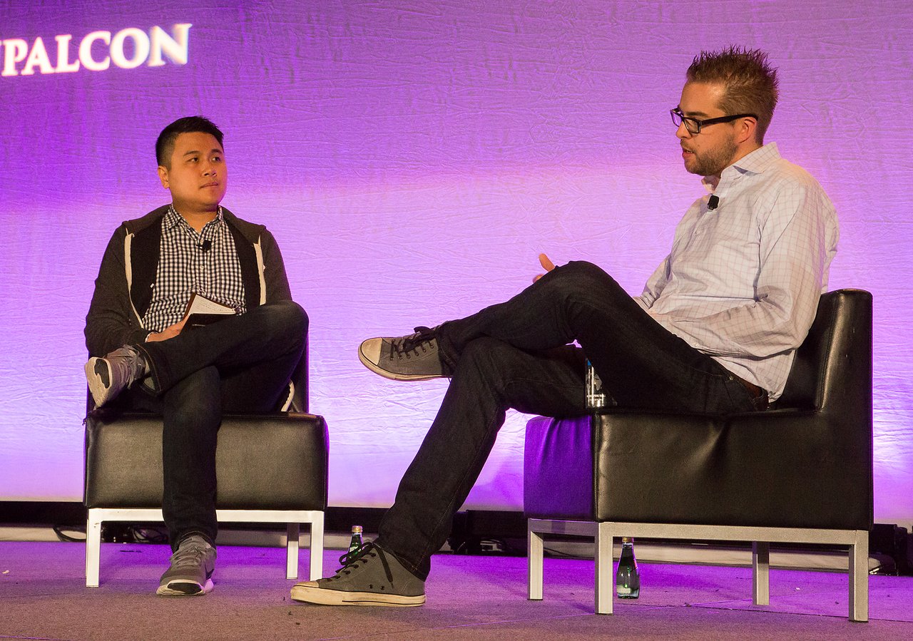 Two people seated on stage in a discussion, one holding a notebook and the other gesturing while speaking.