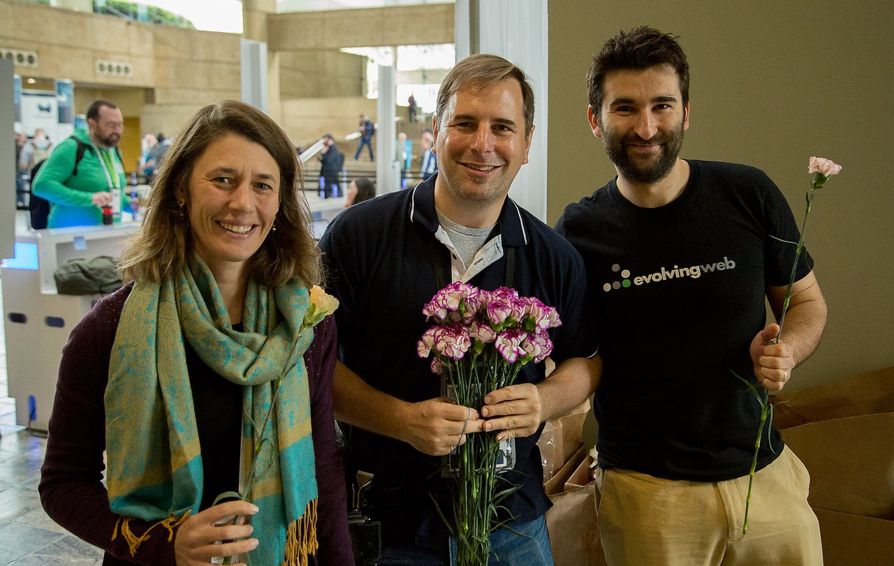Three people smiling while holding pink flowers at an indoor event.