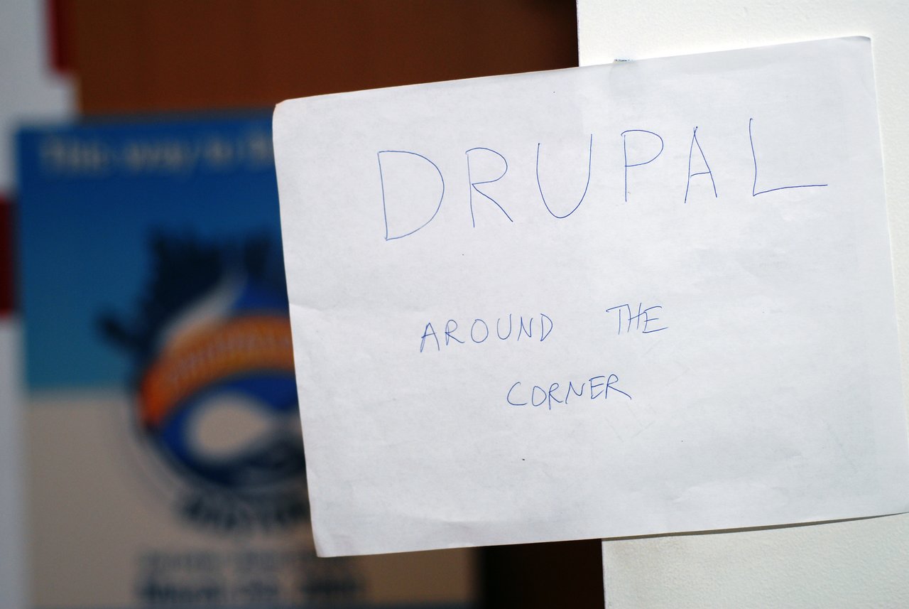 A handwritten sign on a wall reads "Drupal around the corner", directing attendees to a Drupal-related event nearby.