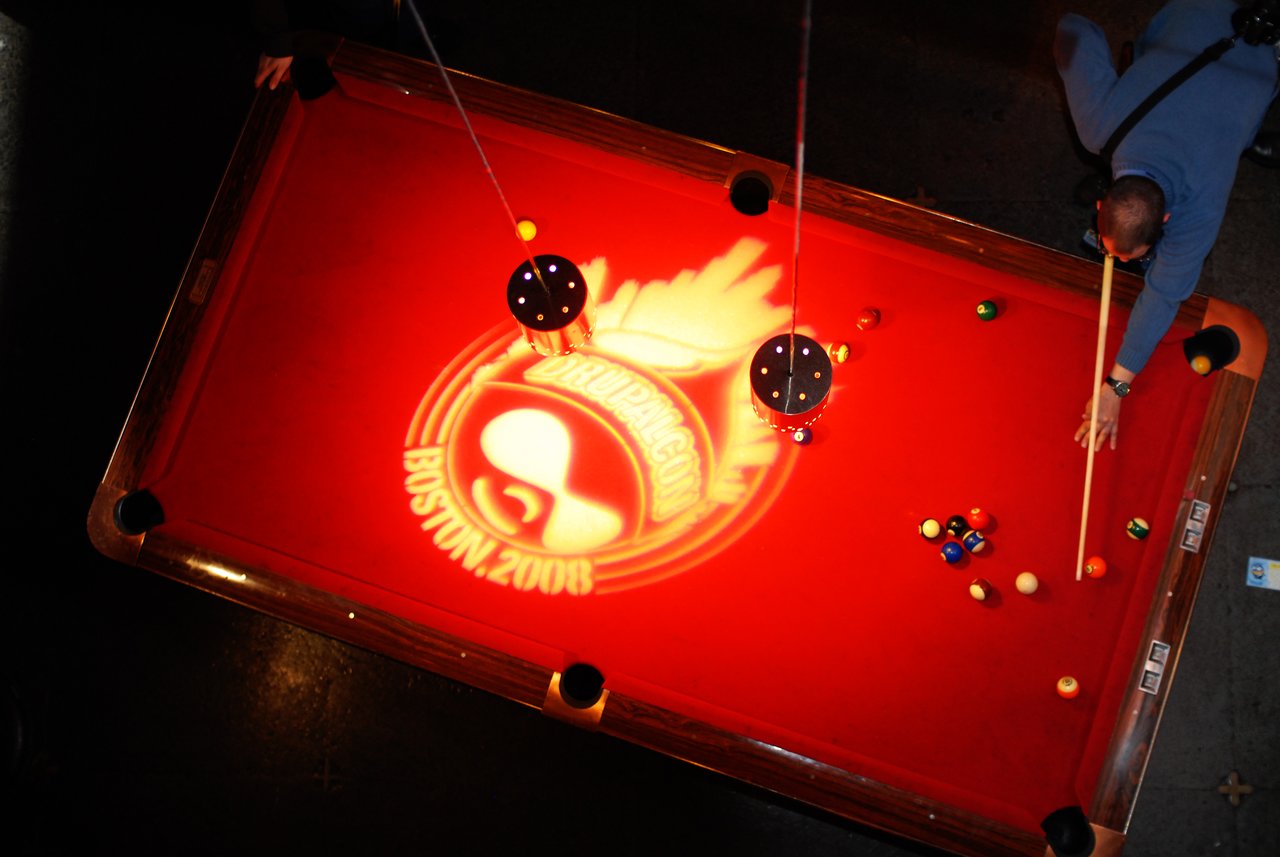 A person plays billiards on a red pool table with a "DrupalCon Boston 2008" logo projected onto it.