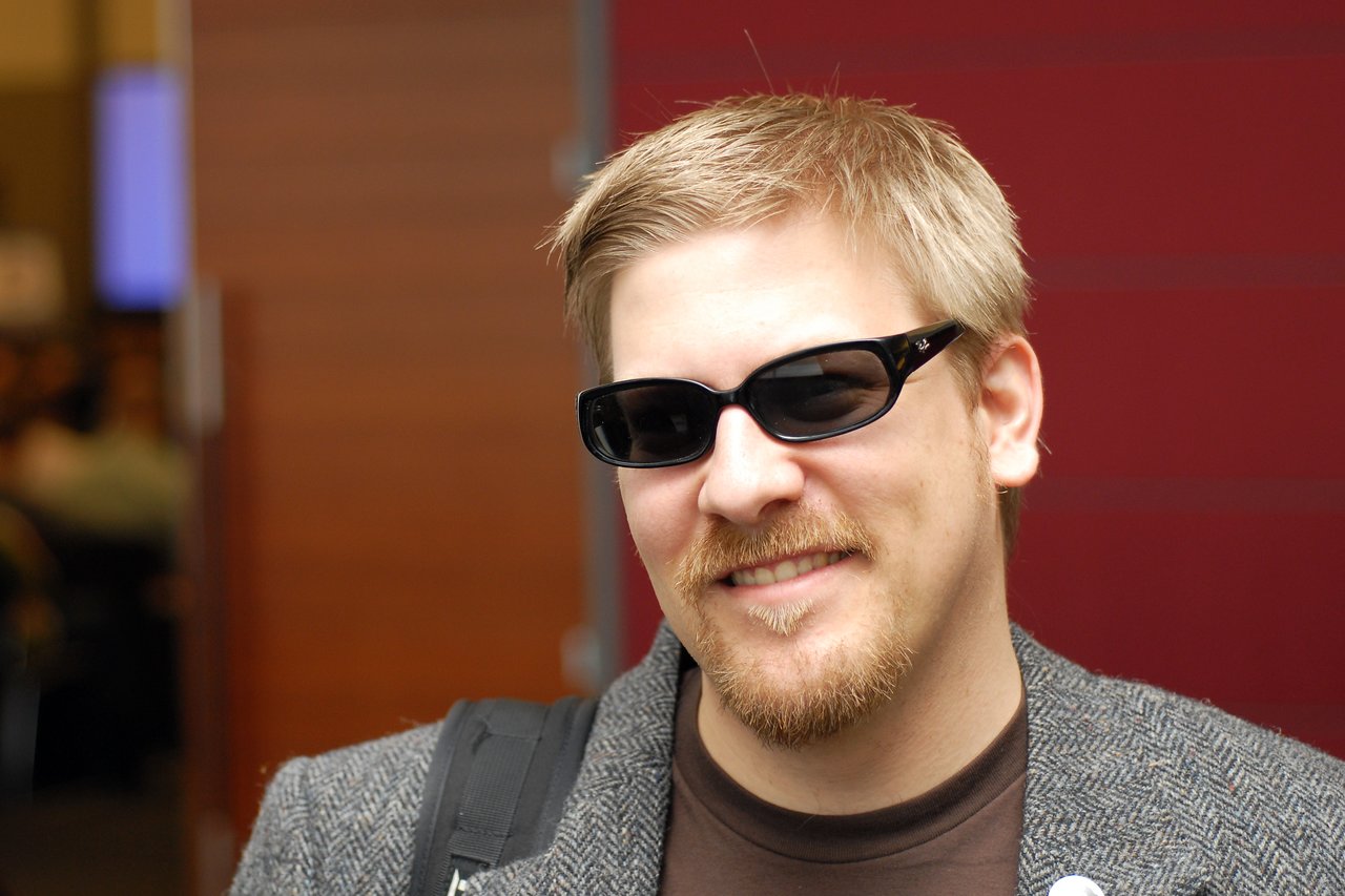 A man with sunglasses and a beard smiles while wearing a backpack at DrupalCon Boston 2008.