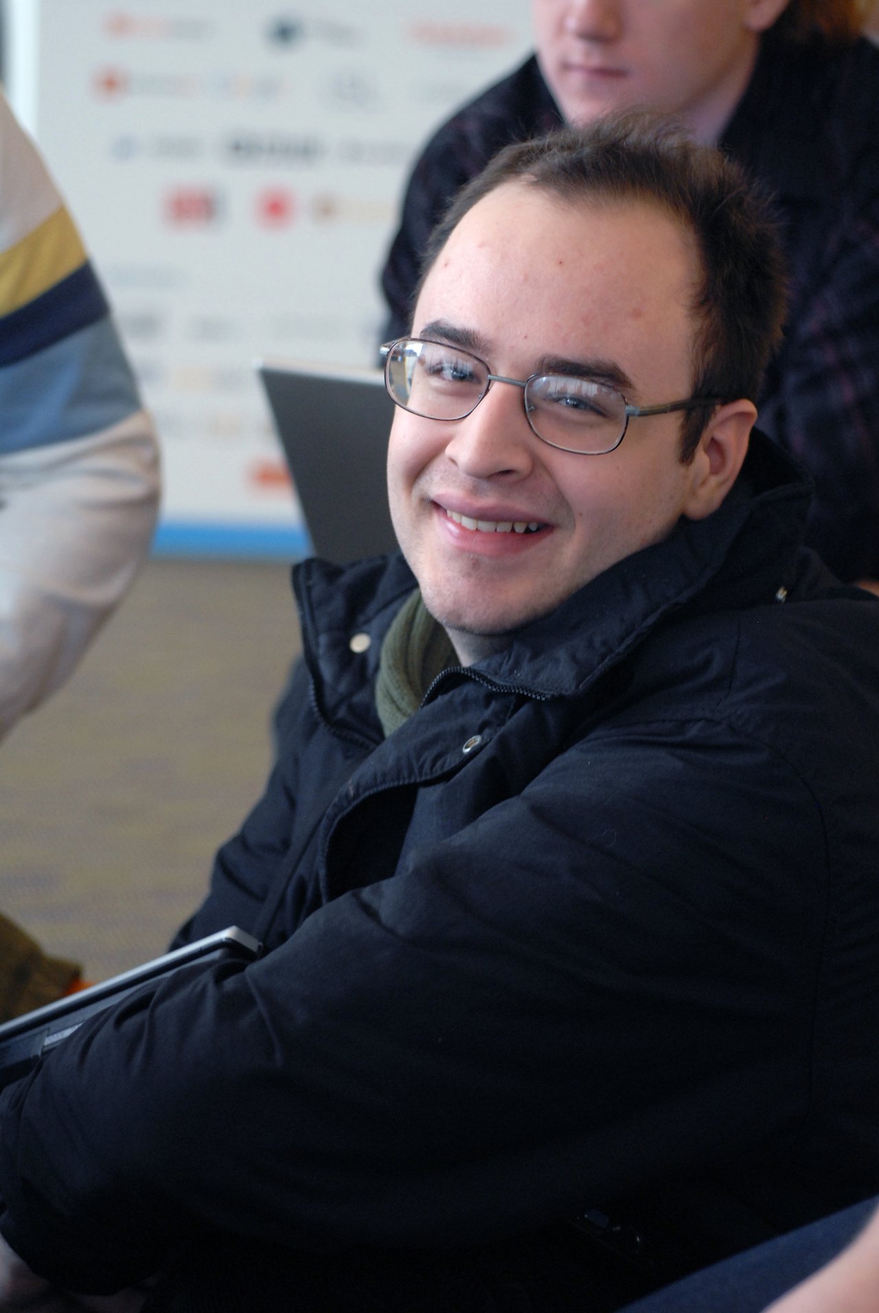 A person wearing glasses and a black jacket smiles at the camera while sitting indoors with a laptop nearby.