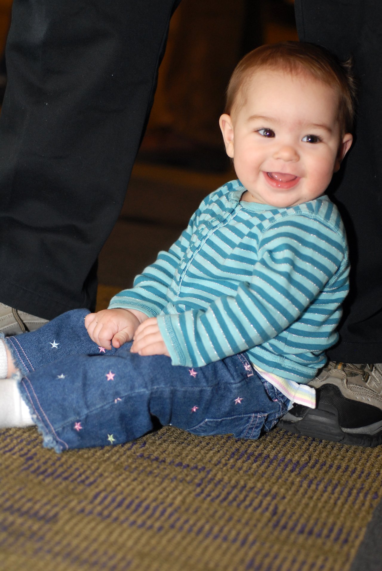 A smiling baby in a striped shirt sits on the floor, leaning against an adult's legs.