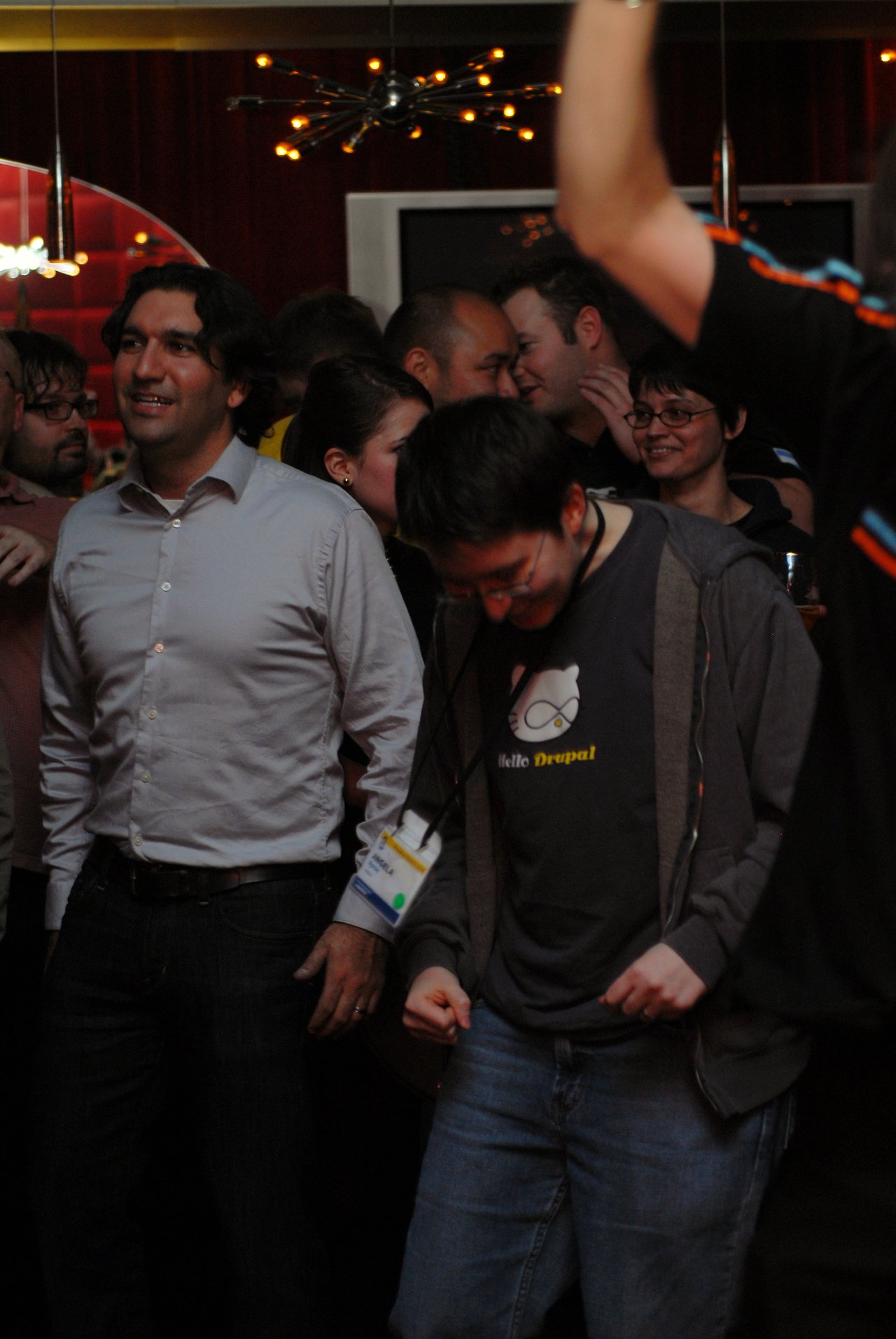 A person in a "Hello Drupal" shirt dances energetically in a crowded room while others socialize and watch.
